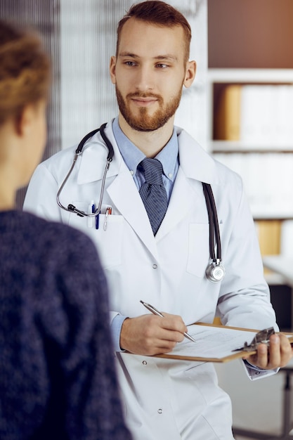 Amichevole medico barbuto e donna paziente che discutono dell'attuale esame sanitario mentre si siedono nella soleggiata clinica. Concetto di medicina