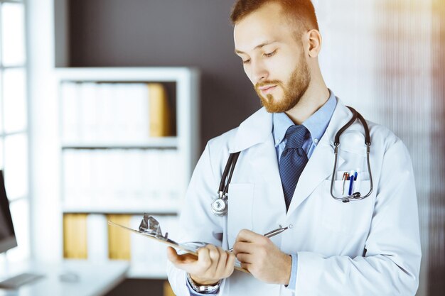 Amichevole medico con barba rossa in piedi e scrivendo con appunti nella clinica soleggiata nel suo posto di lavoro Concetto di medicina