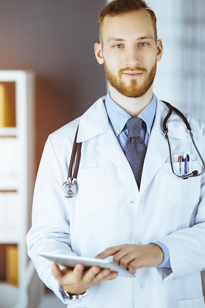 Amichevole medico con la barba rossa che utilizza il computer tablet nella clinica soleggiata nel suo posto di lavoro. Concetto di medicina.