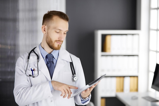 Amichevole medico con la barba rossa che utilizza un tablet in clinica nel suo posto di lavoro. Servizio medico perfetto in ospedale e concetto di medicina.