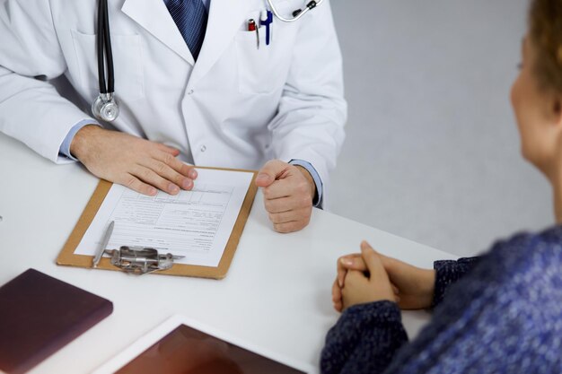 Amichevole medico con la barba rossa e donna paziente che discutono dell'attuale esame sanitario mentre sono seduti in una clinica soleggiata. Concetto di medicina.