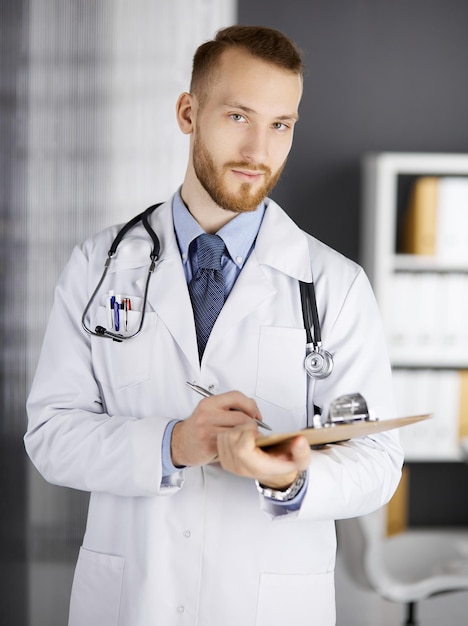 Amichevole medico con la barba rossa in piedi e scrivendo con appunti in clinica al suo posto di lavoro. Servizio medico perfetto in ospedale. Concetto di medicina.