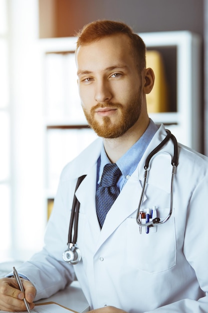 Amichevole medico con la barba rossa seduto e scrivendo negli appunti in clinica soleggiata. Concetto di medicina.