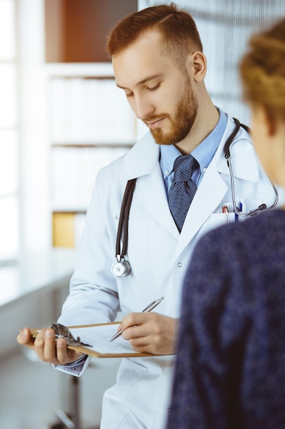 Amichevole medico e paziente con barba rossa che discutono dell'attuale esame sanitario mentre sono seduti in una clinica soleggiata Concetto di medicina