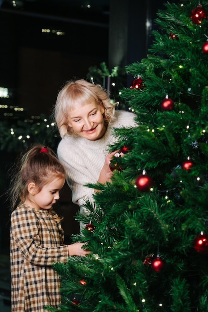 Amichevole nonna e bambina che decorano un albero di natale con palline rosse lucide. al chiuso