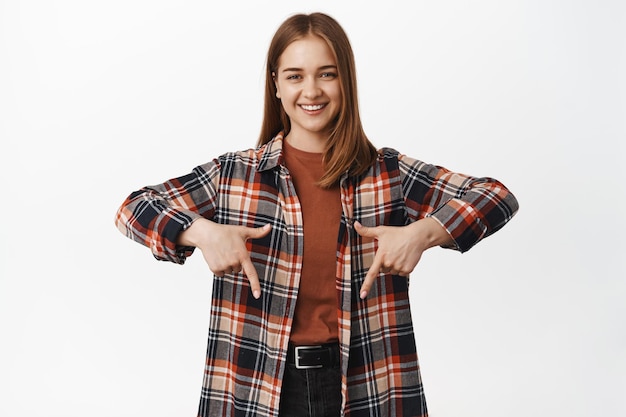 Amichevole ragazza millenaria sorridente, puntando il dito verso il basso, mostrando annuncio, pubblicità o logo, indicando banner, in piedi in abiti casual su sfondo bianco.