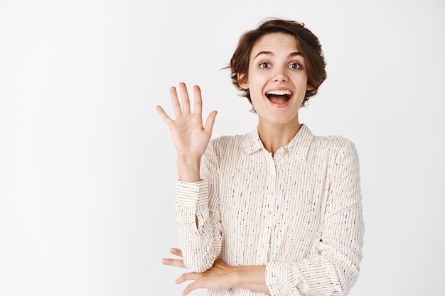 Amichevole ragazza naturale che saluta, agitando la mano e sorridendo gioiosa, salutandoti, in piedi sul muro bianco