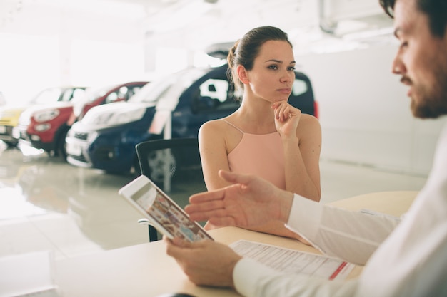 Amichevole venditore di automobili che parla con una giovane donna e mostra una nuova auto all'interno dello showroom Firma del contratto.