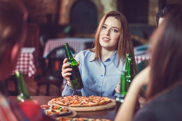 Amici a bere qualcosa in un bar, sono seduti a un tavolo di legno con birre e pizza. Concentrati su una bellissima ragazza che tocca la sua bottiglia.