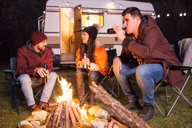 Amici allegri che ridono e bevono birra in un campeggio intorno al fuoco. Camper retrò.