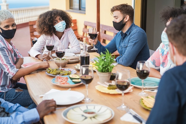 Amici che mangiano e bevono vino insieme a maschere protettive