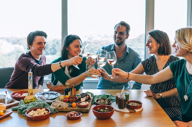 Amici che salgono bicchieri di vino per un brindisi celebrativo a casa seduti a un tavolo da festa pieno di finger food