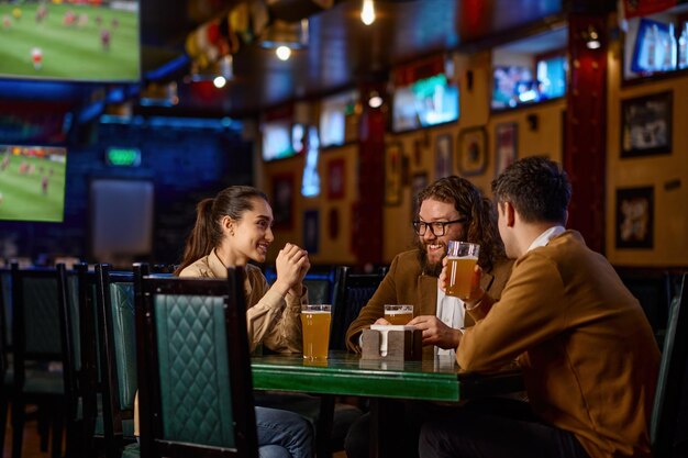 Amici che si divertono seduti al tavolo del pub. Giovani che parlano bevendo birra e utilizzando lo smartphone