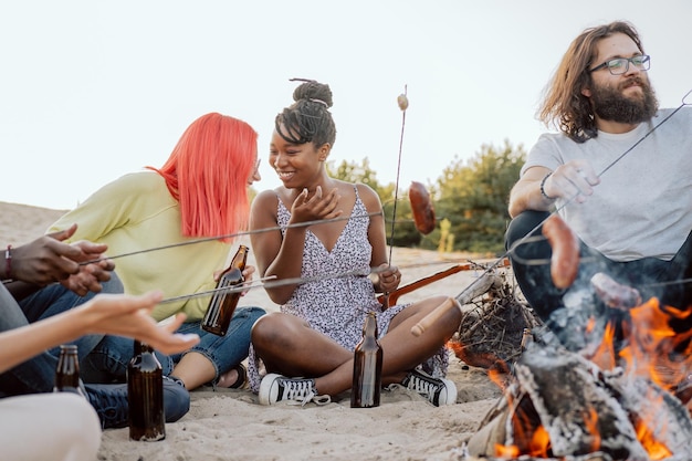 Amici di diverse nazionalità sono seduti sulla spiaggia vicino al fuoco sulla sabbia a bere birra