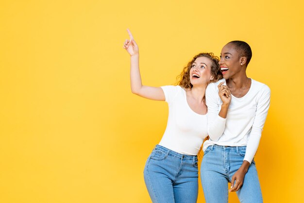 Amici di donna interrazziale felici gioiosi che puntano e cercano lo spazio vuoto su sfondo giallo isolato per studio