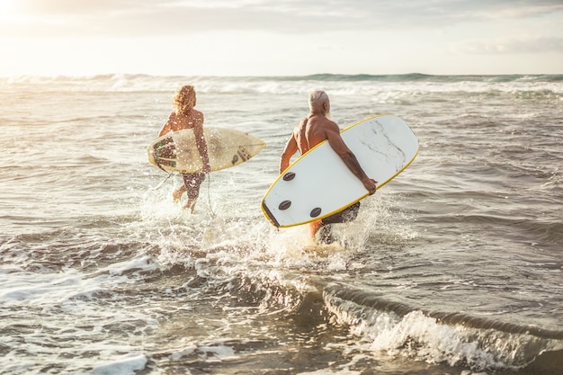 Amici di età diverse in esecuzione sulla spiaggia al tramonto per l'allenamento di surf