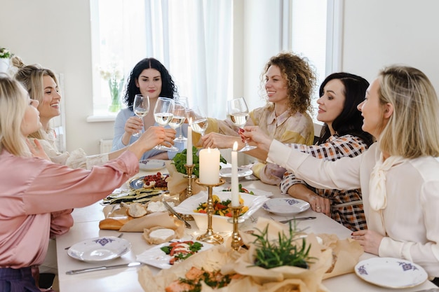 Amici di giovani donne felici che tintinnano i bicchieri e sorridono al tavolo festivo servito con cibo e vino bianco decorato con candele