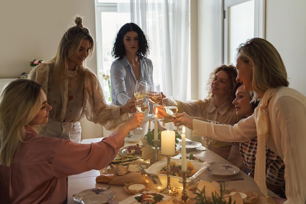 Amici di giovani donne felici che tintinnano i bicchieri e sorridono al tavolo festivo servito con cibo e vino bianco decorato con candele