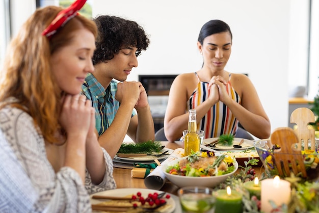 Amici diversi e felici seduti a tavola e pregando prima di cena a Natale