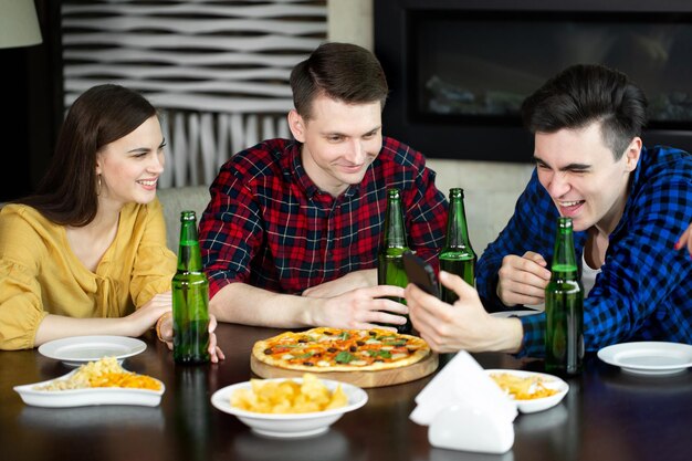 Amici felici guardano il telefono mentre bevono birra e pizza in un bar