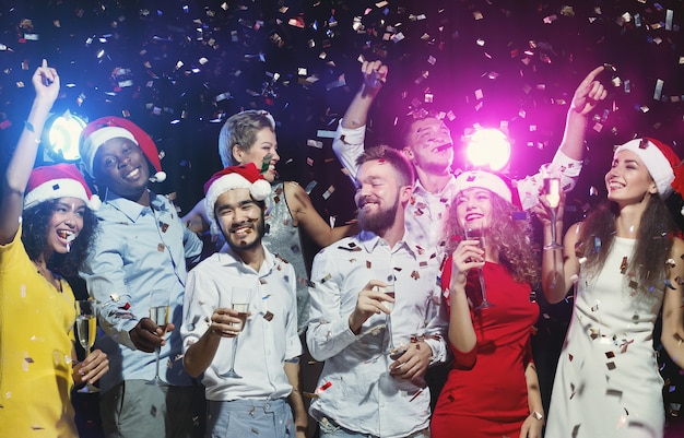 Amici felici in cappelli di Babbo Natale che ballano alla festa di Capodanno sotto i coriandoli