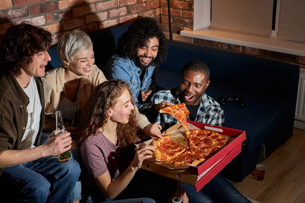 Amici giovani e felici che mangiano pizza e guardano film o serie tv a casa, gli studenti americani si godono il tempo libero dopo le lezioni, riposandosi dopo una dura settimana