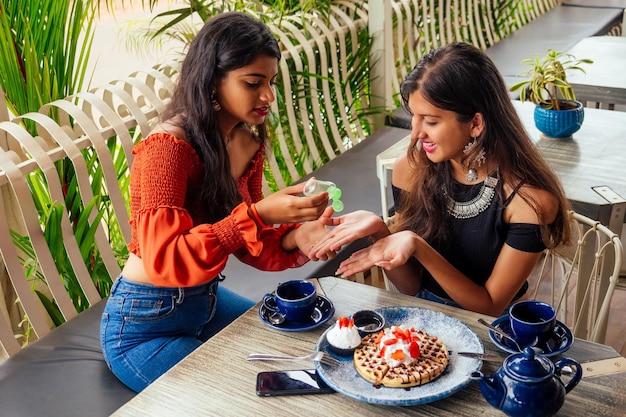 Amici indiani alla moda coppia di due donne che usano gel antibatterico su amici che hanno atteso da tempo l'incontro con gli studenti di gossip che mangiano pancake bevendo tè masala dopo lo shoppingsummer stile di vita instahram