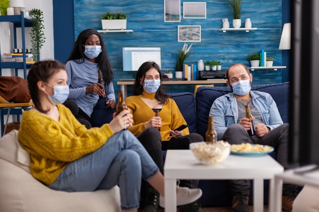 Amici multietnici che guardano la televisione seduti sul divano a bere birra indossando la maschera facciale durante l'epidemia di pandemia di covid come prevenzione contro la diffusione del virus, divertendosi nel tempo libero.