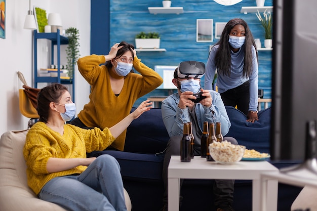 Amici multietnici che si godono la tecnologia vr giocando ai videogiochi in soggiorno indossando una maschera per prevenire l'infezione da coronavirus mantenendo le distanze sociali. Diverse persone che si divertono alla nuova festa normale