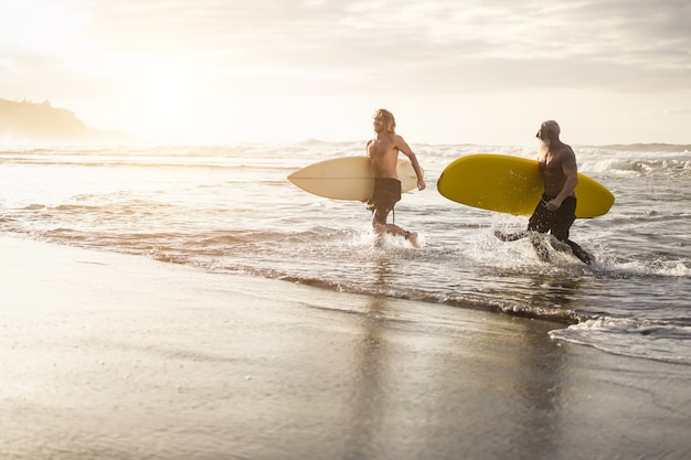 Amici multigenerazionali che corrono lungo la spiaggia con le tavole da surf sulla spiaggia dell'isola - Focus sui volti