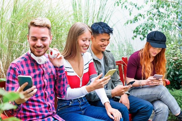 Amici multirazziali sorridenti che usano gli smartphone sulla panchina