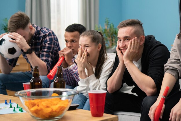 Amici sconvolti che guardano il calcio e bevono a casa seduti su un divano Studenti che guardano lo sport su t