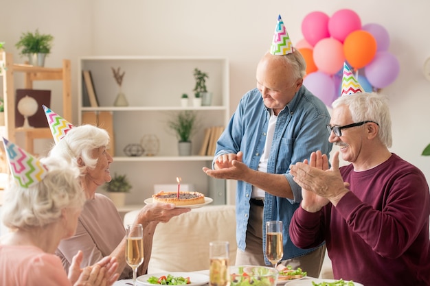 Amici senior che applaudono le mani mentre guardano il loro amico con la torta di compleanno dal tavolo servito