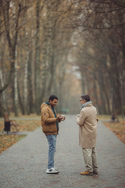 Amici un anziano e un giovane che camminano, parlano e bevono caffè insieme nel parkxA autunnale