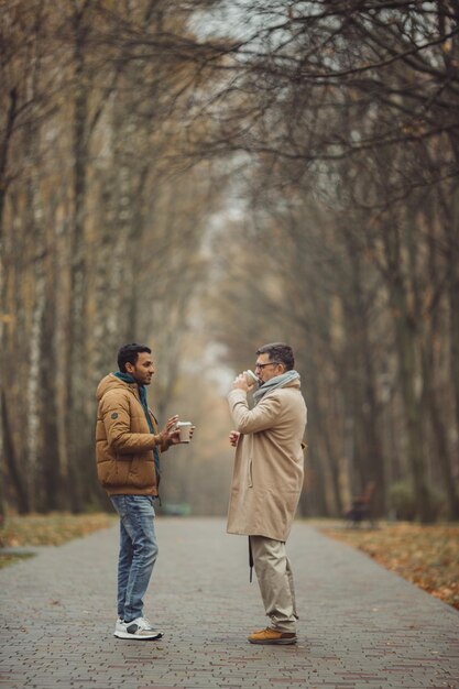 Amici un anziano e un giovane che camminano, parlano e bevono caffè insieme nel parkxA autunnale