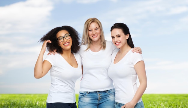 amicizia, diversità, corpo positivo e concetto di persone - gruppo di donne felici di diverse dimensioni in magliette bianche che si abbracciano sopra il cielo blu e lo sfondo dell'erba