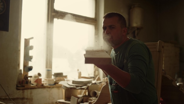 Amico in primo piano che soffia su una tavola di legno in un laboratorio di falegnameria Serio artigiano che soffia la segatura in studio Ritratto di un falegname concentrato che pulisce il legno in casa