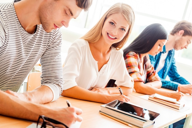 Amo studiare! Sorridente giovane donna che guarda la telecamera e sorride mentre i suoi compagni di classe scrivono qualcosa