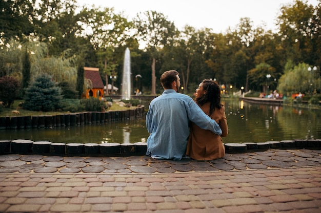 Amore coppia che abbraccia allo stagno nel parco estivo. L'uomo e la donna si rilassano all'aperto, prato verde. Famiglia che abbraccia vicino al lago in estate, weekend nella natura