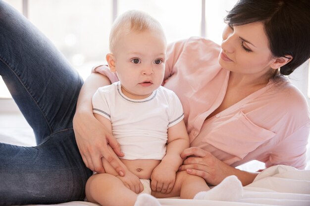 Amore materno. Bella giovane madre sdraiata sul lato del letto mentre il piccolo bambino si siede vicino a lei