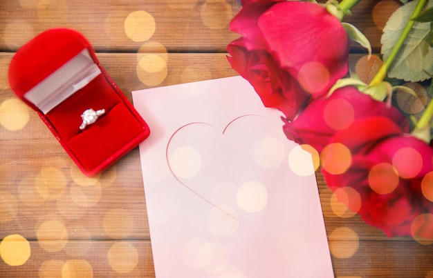 amore, proposta, giorno di san valentino e concetto di vacanze - primo piano della confezione regalo con anello di fidanzamento con diamante, rose rosse e biglietto di auguri su legno