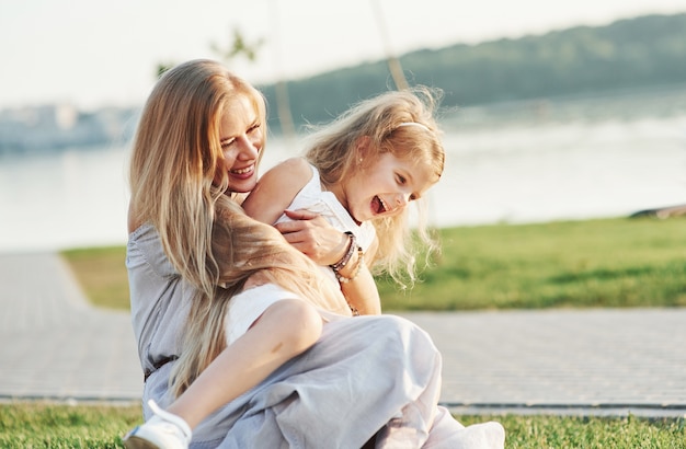 Amore puro. Foto di giovane madre e sua figlia che si divertono sull'erba verde con il lago sullo sfondo.