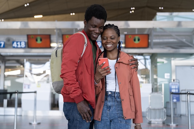 Amorevole coppia di viaggiatori africani in attesa del volo nel terminal dell'aeroporto guardando il telefono