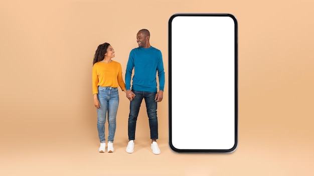 Amorevole coppia nera in piedi vicino al grande smartphone con schermo vuoto che si tiene per mano mockup colpo a figura intera