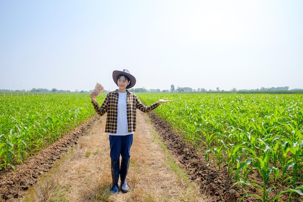 Ampio angolo di agricoltore asiatico donna indossare camicia in piedi e tenendo banconota thailandese alla fattoria di mais verde