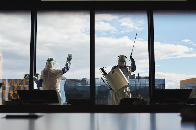 Ampio angolo vista posteriore ritratto di due lavoratori che indossano tute ignifughe disinfezione di windows in edificio per uffici in piedi contro il cielo blu,