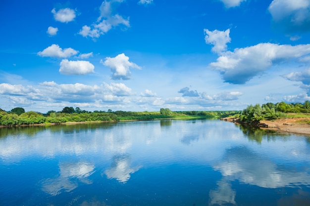 Ampio fiume con riflessione e cespuglio verde sulle coste e cielo nuvoloso blu