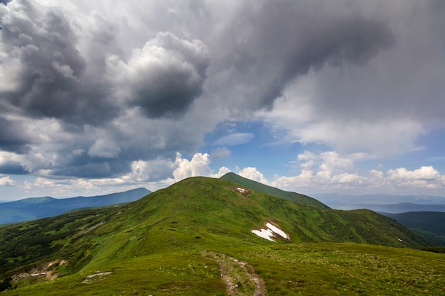 Ampio panorama illuminato dalla valle verde del sole del mattino, colline ricoperte di foresta e lontane montagne nebbiose con macchie di neve sotto il cielo ventoso nuvoloso. Bellezza della natura, turismo e concetto di viaggio.