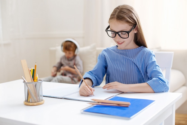 Analitico ambizioso bambino dotato seduto al tavolo e lavorando sul suo compito a casa mentre suo fratello gioca con il tablet su un divano dietro di lei