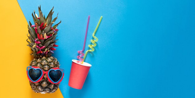 Ananas in occhiali da sole a forma di cuore e tazza di plastica con cannucce su sfondo mezzo blu e giallo